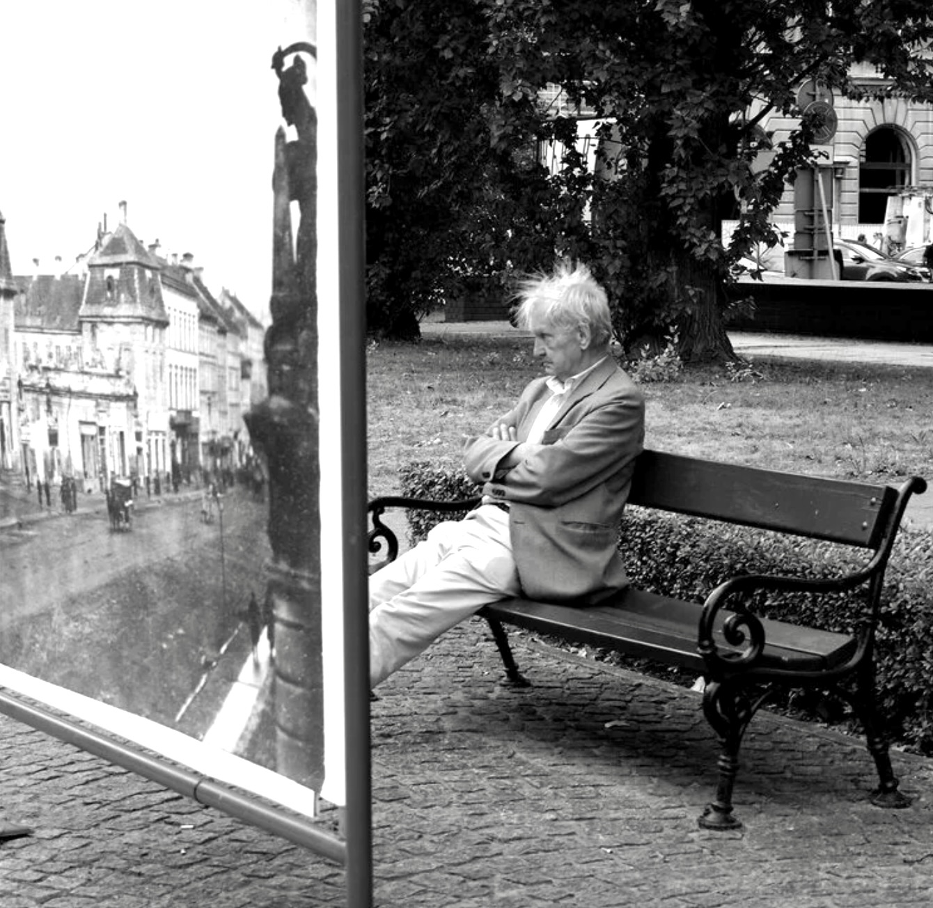 Thinking, Warsaw — a man sits on a bench beside a large vintage photograph of a European street.