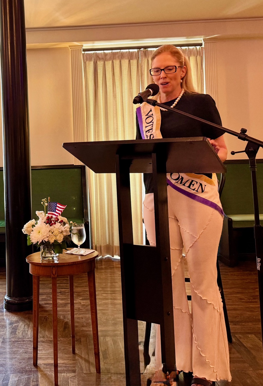 Cynthia Safford-Mulcahy at the podium wearing a 'Votes for Women' sash.