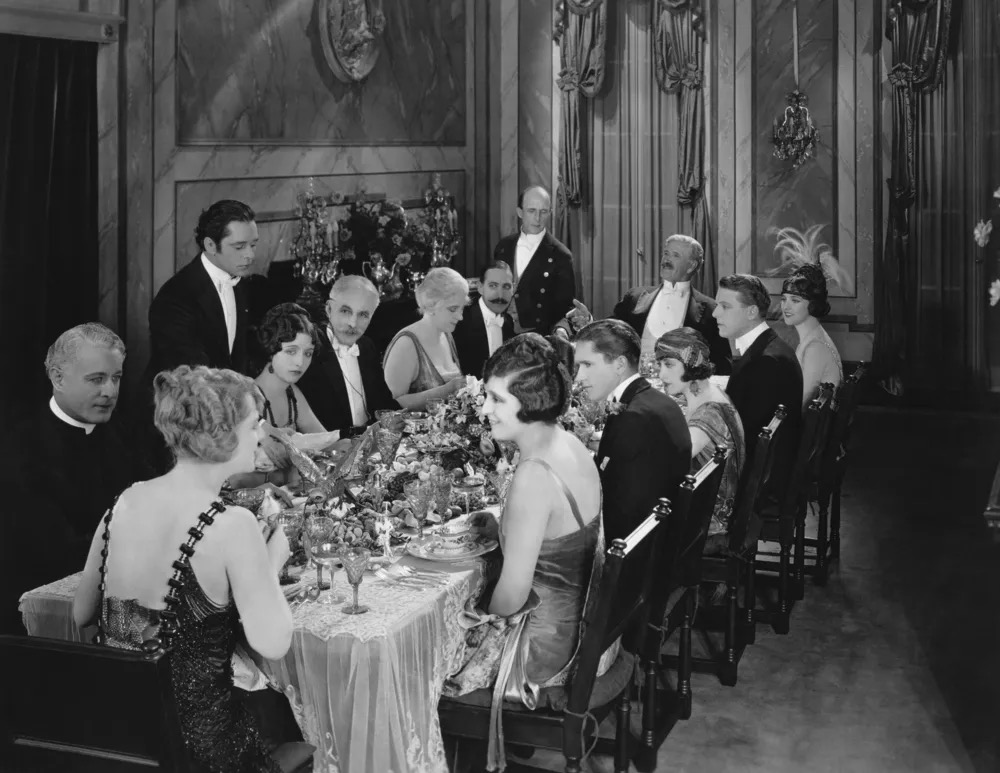 A 1920s formal dinner party, guests in evening dress around an elegantly set table with butlers in attendance
